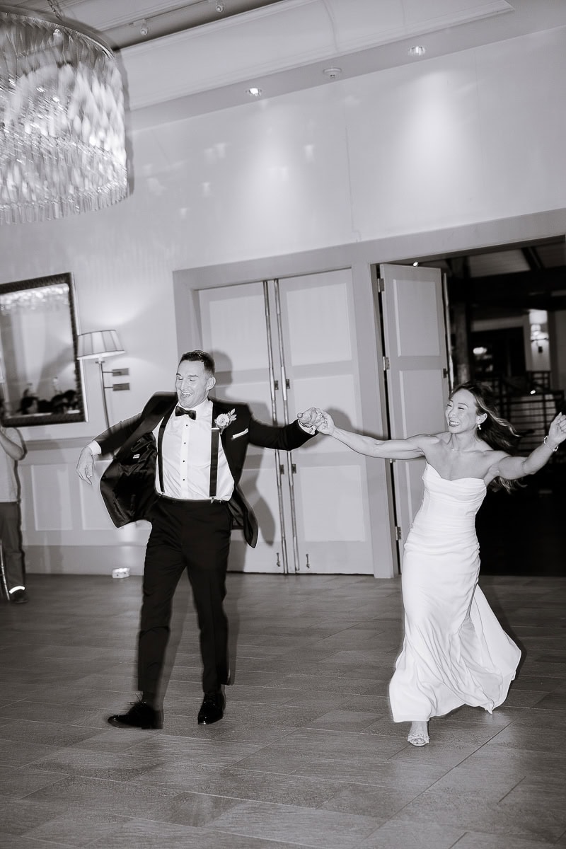 Bride and groom holding hands and dancing energetically in a spacious, elegantly decorated room with soft lighting and a large chandelier at The Stone House, Warren, NJ.