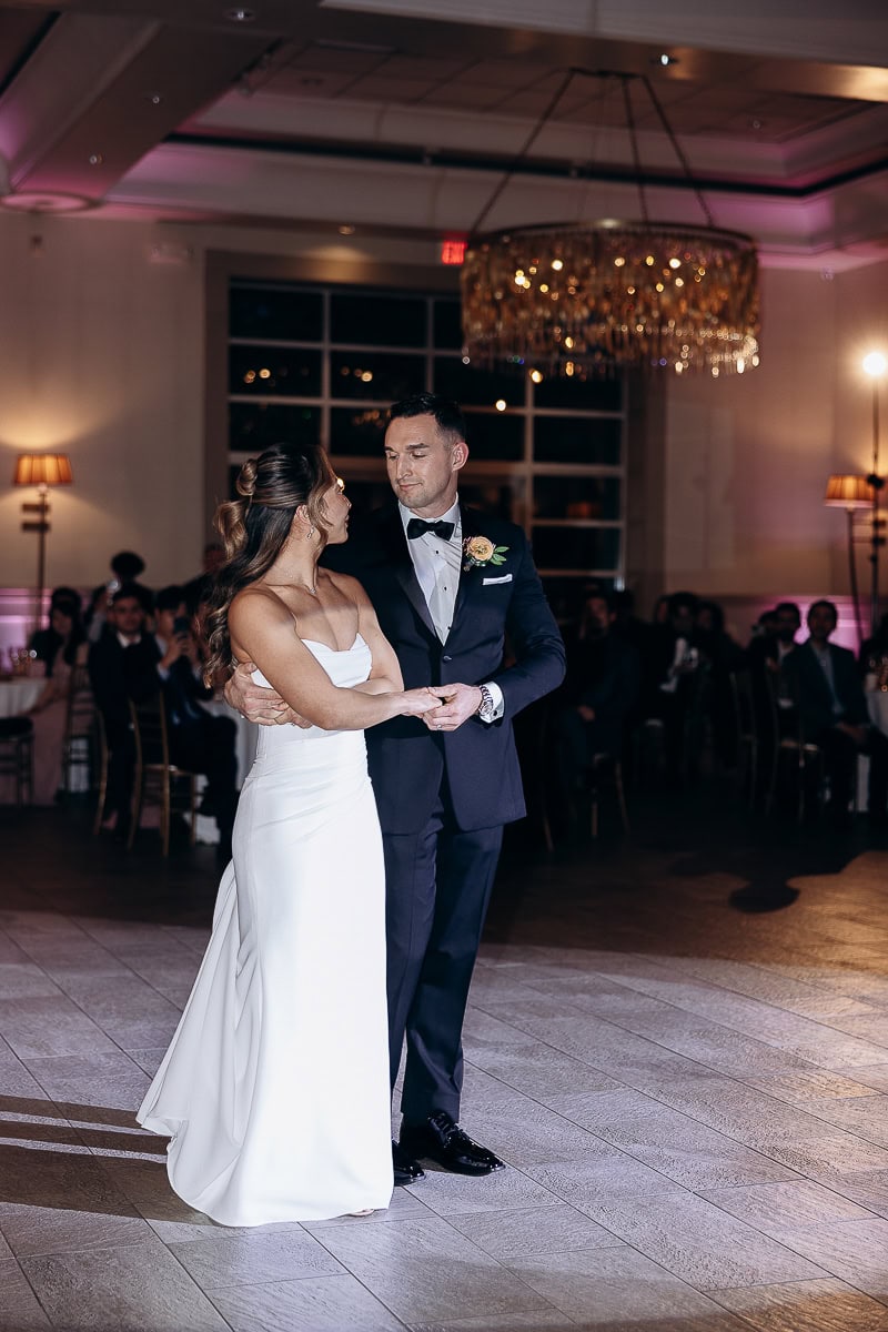 Bride and groom sharing their first dance in an elegant ballroom with a crystal chandelier and seated guests in the background at The Stone House, Warren, NJ.