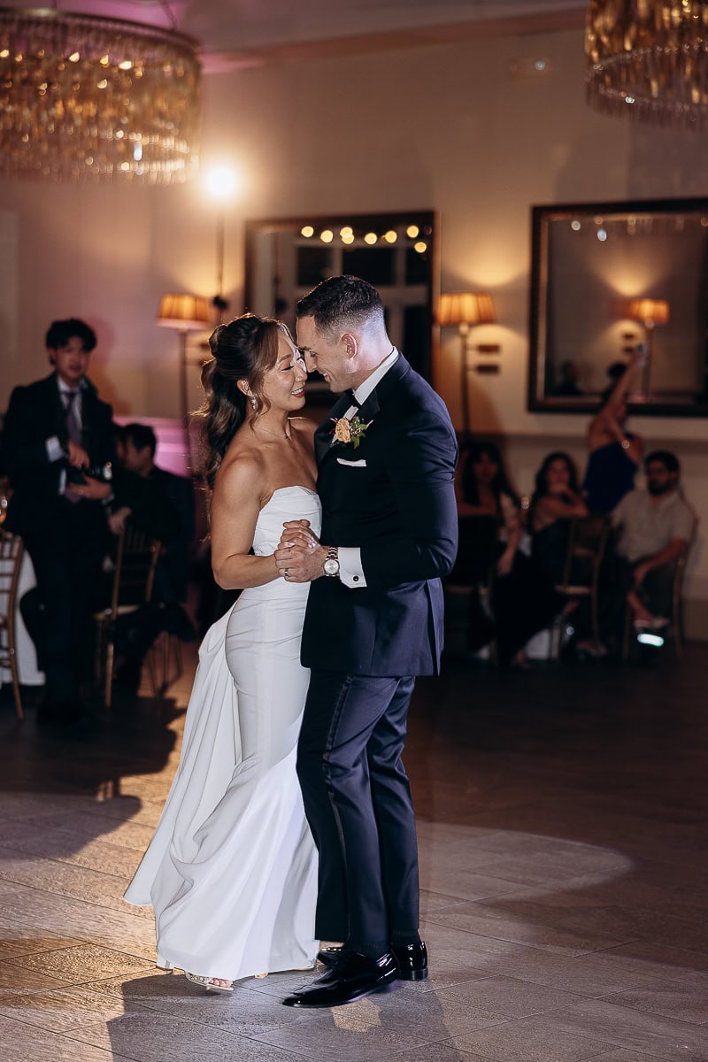 Bride in strapless white gown and groom in black tuxedo dancing closely during first dance at wedding reception with guests seated in background