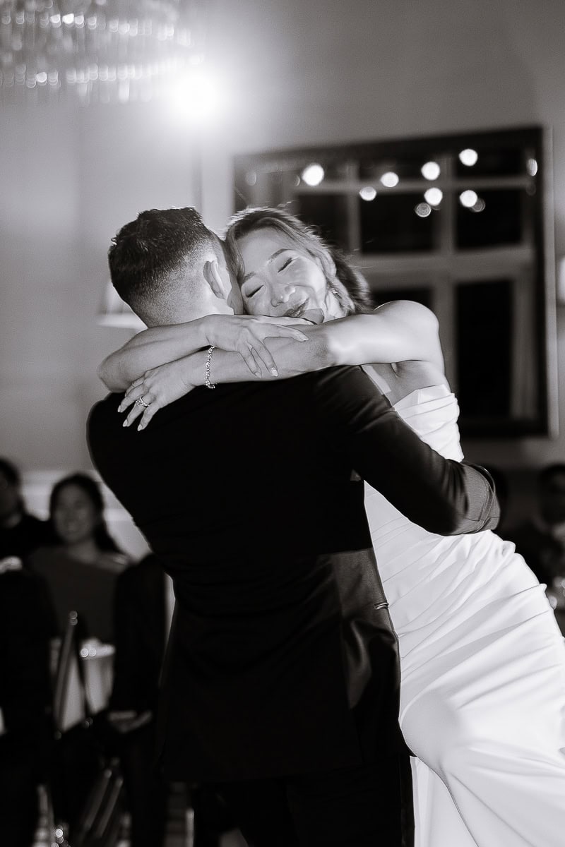 Bride in white gown embraces groom in black suit during wedding dance at The Stone House, Warren, NJ, with soft lighting and blurred guests in background.