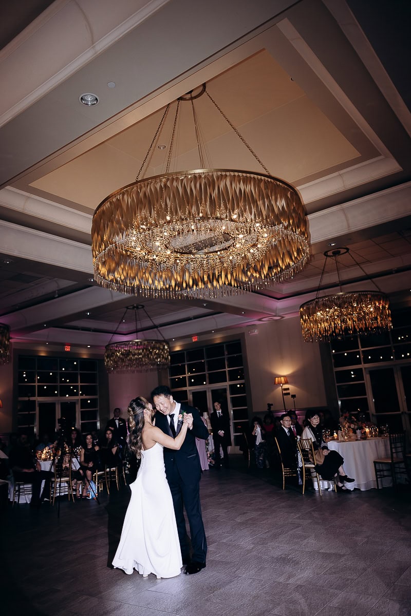 Bride and groom dancing closely under a large ornate gold chandelier in a dimly lit ballroom with seated guests watching.