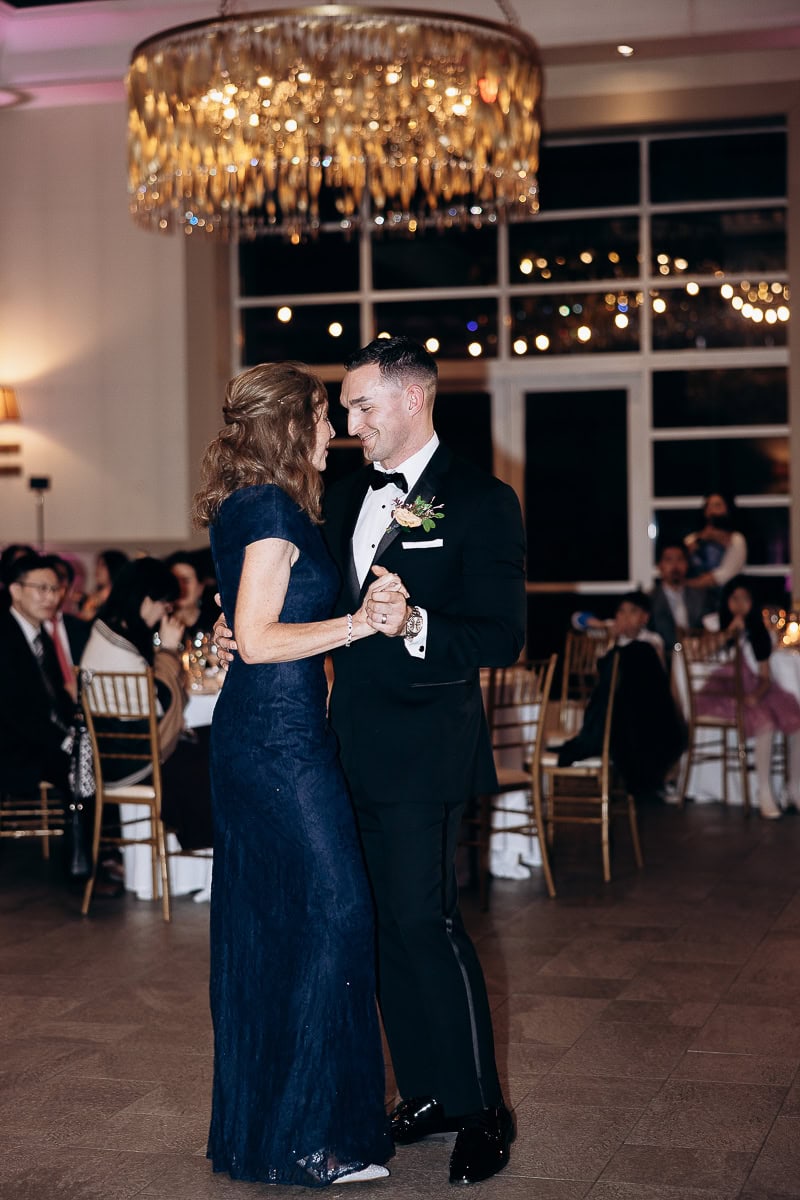 Groom in black tuxedo dancing with mother in navy lace gown beneath crystal chandelier at wedding reception