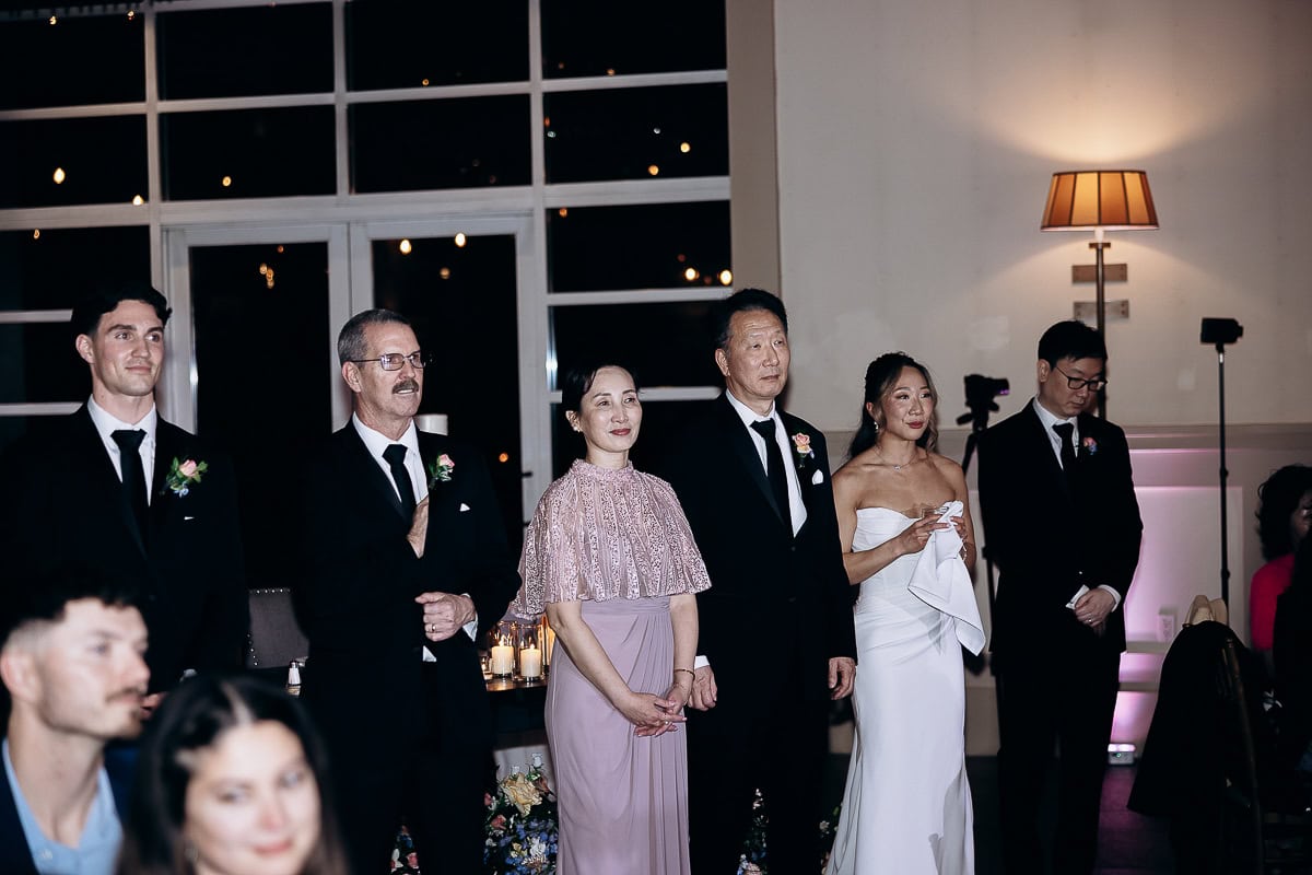 Group of elegantly dressed individuals standing indoors at an evening wedding ceremony with soft lighting and floral arrangements at The Stone House, Warren, NJ.