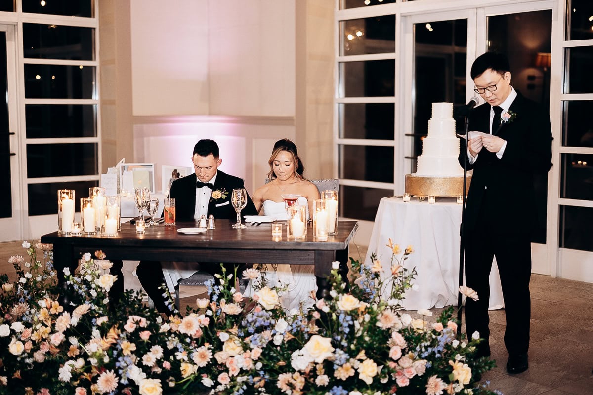 Wedding reception at The Stone House, Warren NJ, with couple seated at a candlelit table surrounded by flowers, and a man giving a speech next to a tiered cake.
