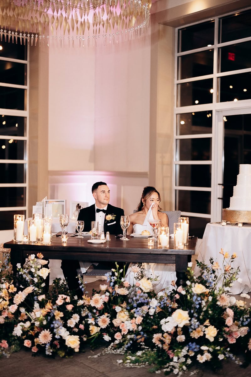 Bride and groom seated at a dark wooden sweetheart table adorned with candles and surrounded by pastel floral arrangements during their wedding reception at The Stone House, Warren, NJ.