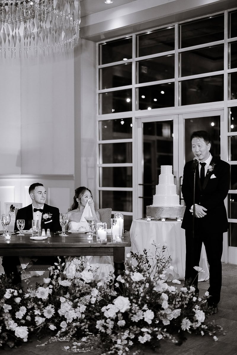 Black and white image of a wedding speech with a man speaking into a microphone beside a tiered cake, while bride and groom sit at a decorated table with flowers and candles.