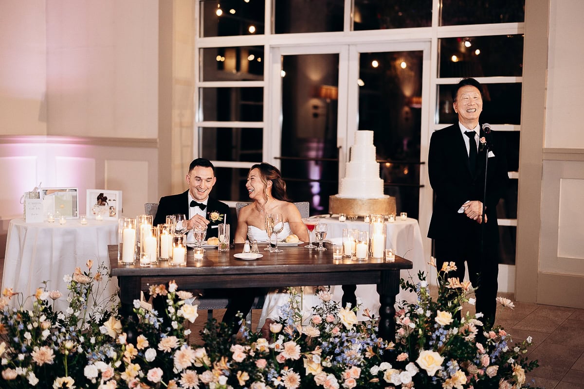 Couple seated at a candlelit table surrounded by flowers during a wedding toast, with a man speaking at a microphone near a tiered cake at The Stone House, Warren, NJ.