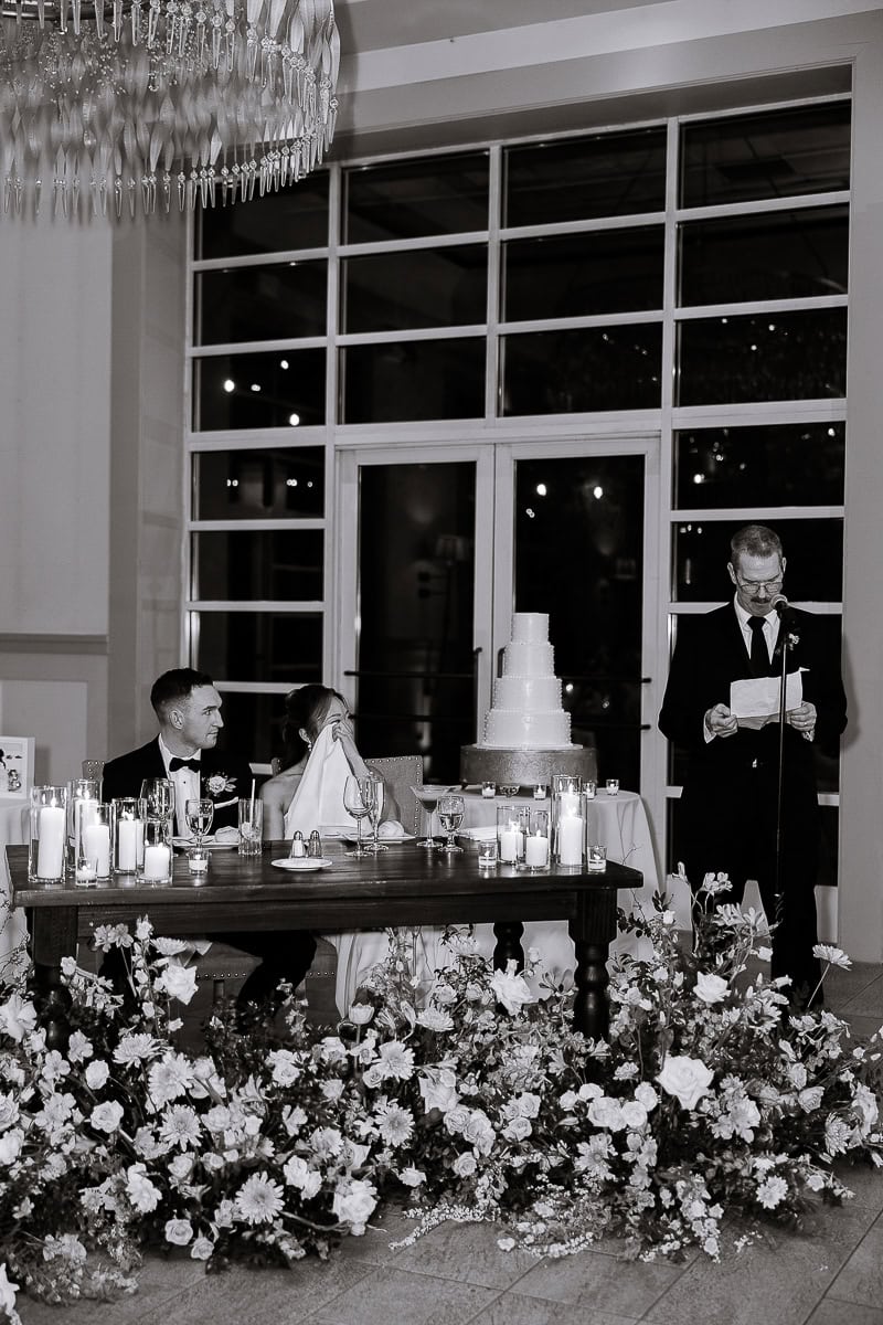 Black and white image of a wedding speech at The Stone House, Warren NJ; bride and groom seated at a floral-adorned table with candles, man reading from paper at microphone.