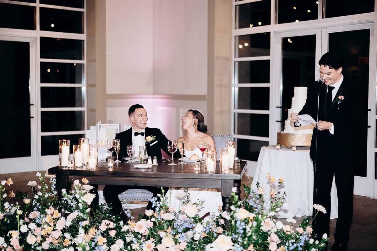 Bride and groom seated at a candlelit table adorned with flowers, smiling as a man in a black suit delivers a speech beside a tiered wedding cake in an elegant venue.