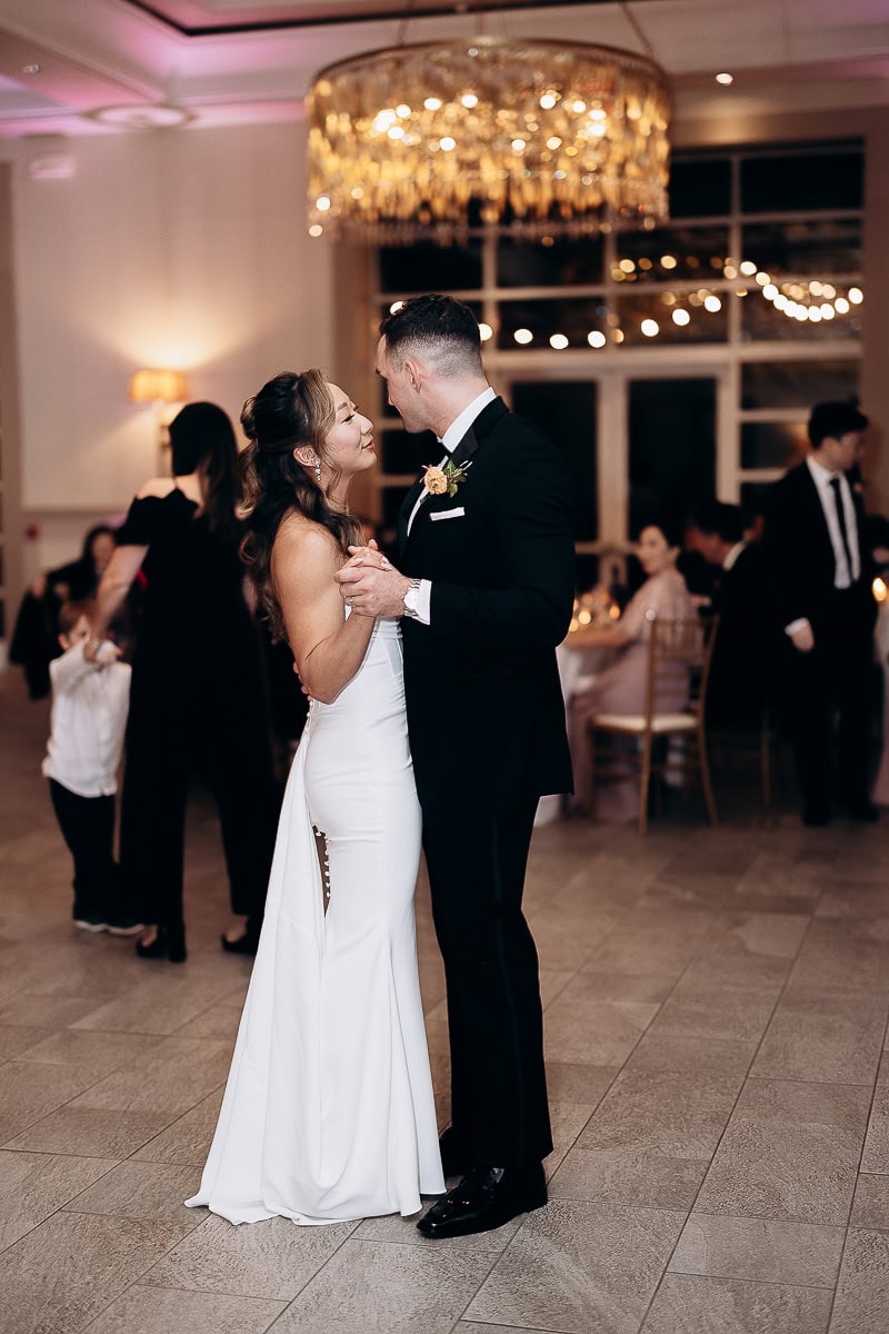 Bride in white gown and groom in black tuxedo dancing closely under a crystal chandelier at an indoor wedding reception at The Stone House, Warren, NJ.