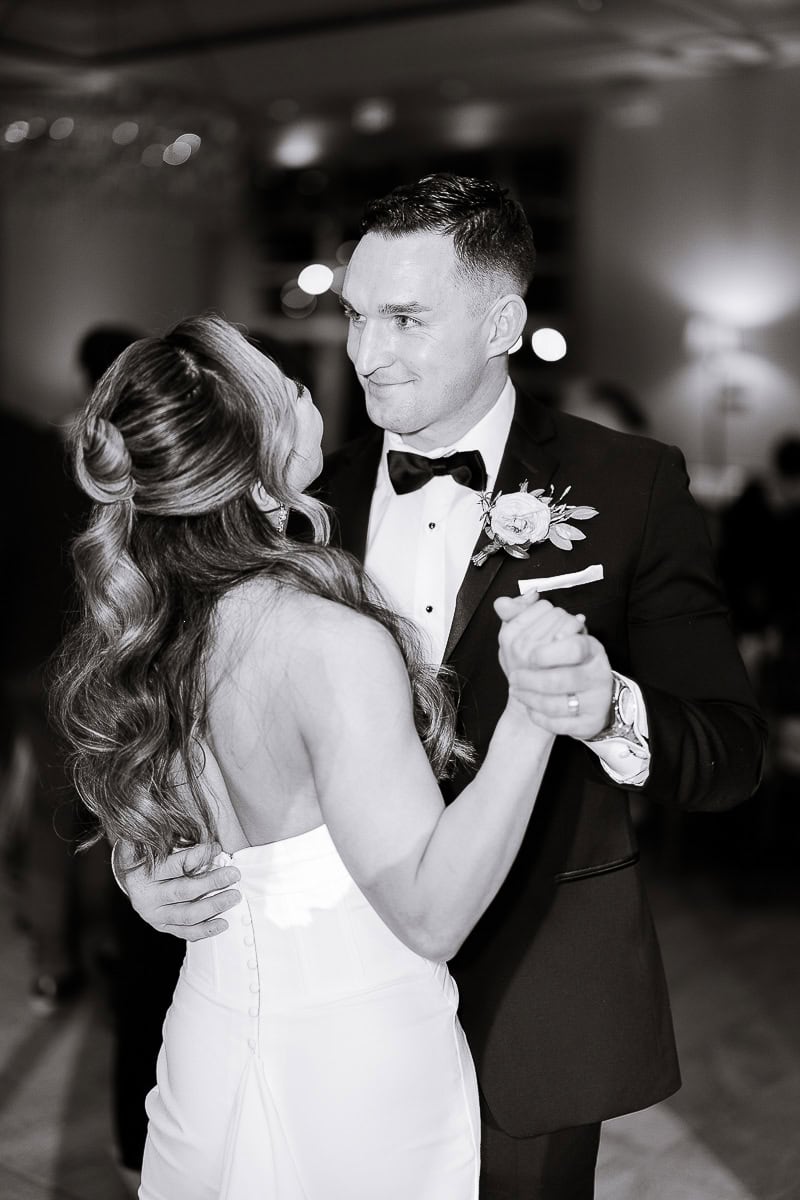 Black and white photo of a couple dancing closely, the groom in a tuxedo with boutonniere and the bride in a strapless dress with long wavy hair, at an indoor wedding venue.