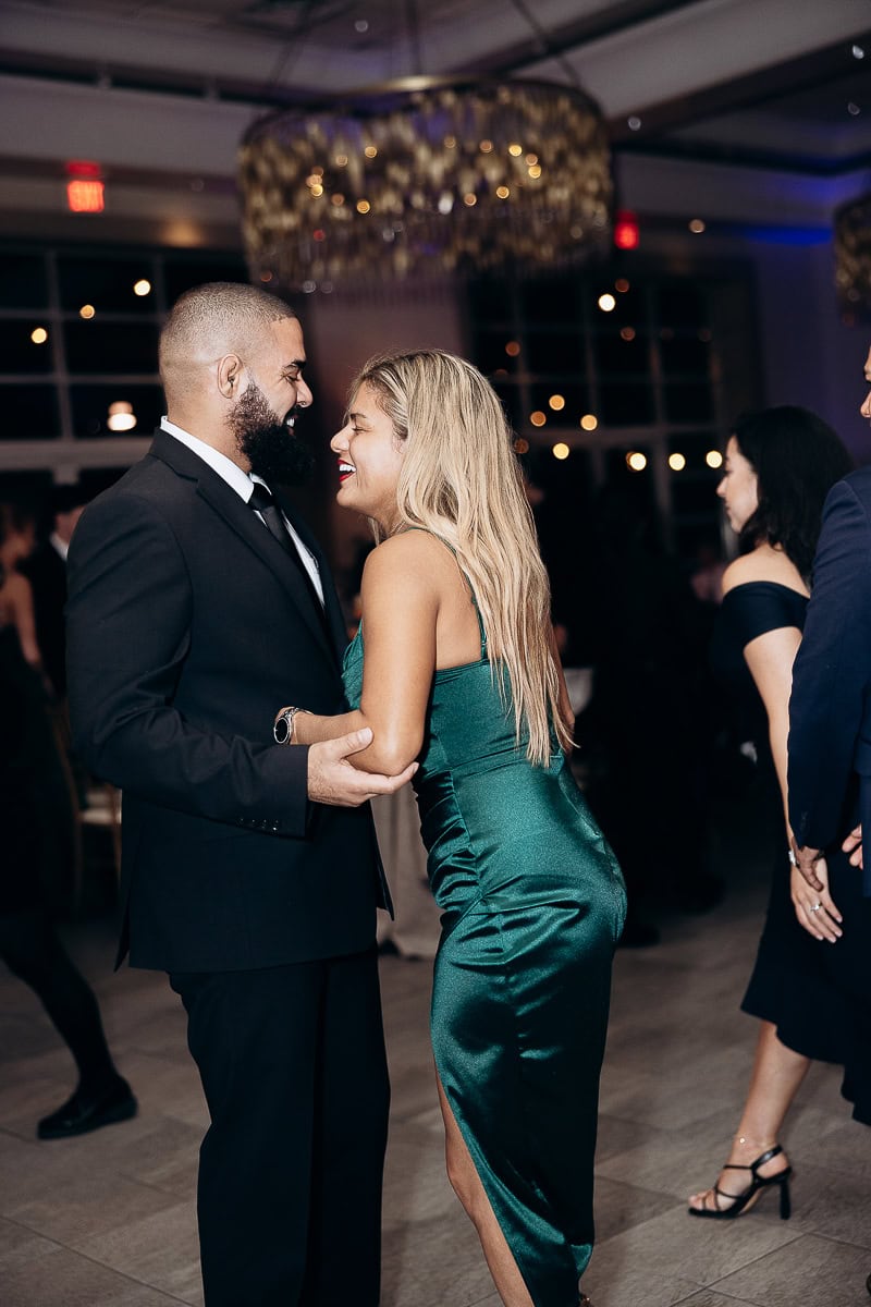 Man in black suit and woman in emerald satin dress share a joyful moment on a dance floor at an elegant indoor wedding venue.