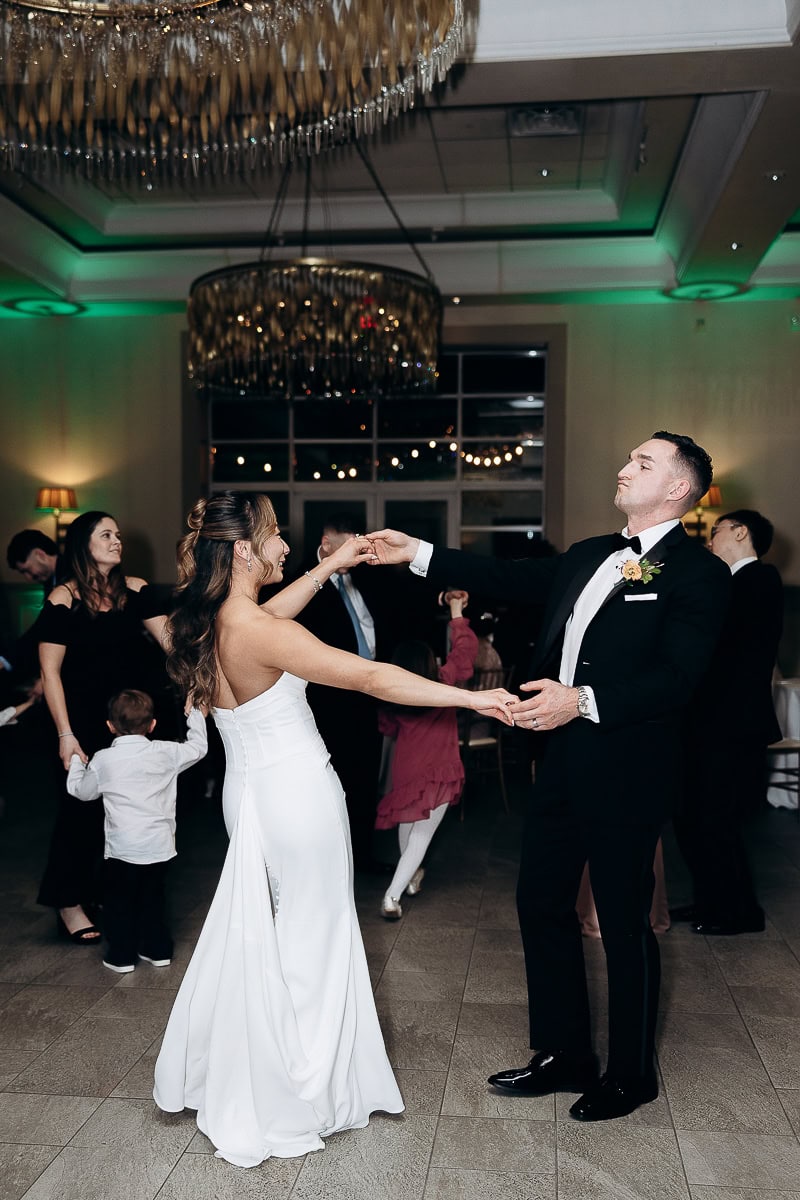 Bride in sleek white gown and groom in black tuxedo dancing together in elegant reception hall with chandeliers at The Stone House, Warren, NJ.