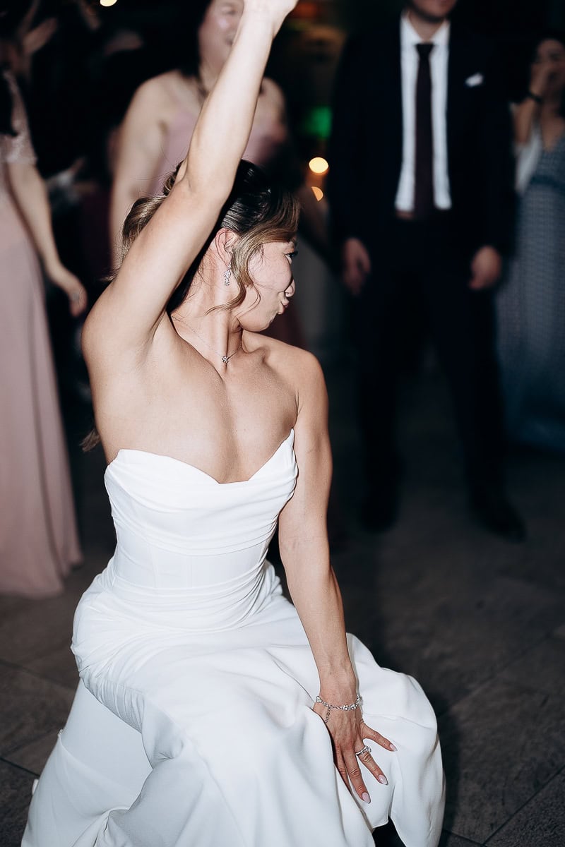 Bride in strapless white wedding dress dancing with arm raised, surrounded by blurred guests at The Stone House, Warren, NJ.