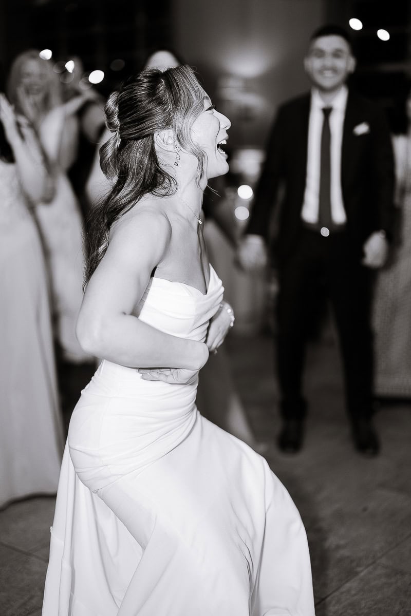 Bride in strapless white gown laughing and dancing, with blurred guests in background at evening wedding reception at The Stone House, Warren, NJ.