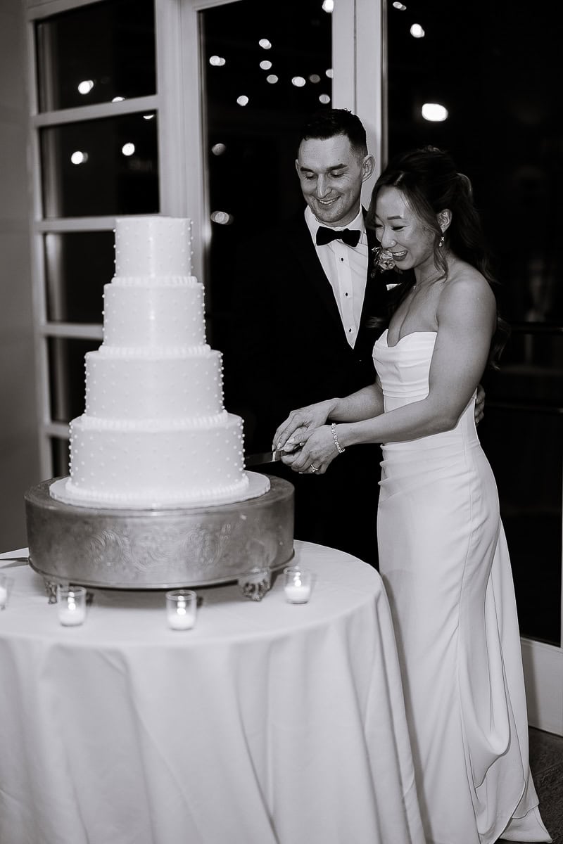Bride and groom cutting a four-tier white wedding cake on a silver stand, surrounded by small candles, at The Stone House, Warren, NJ.