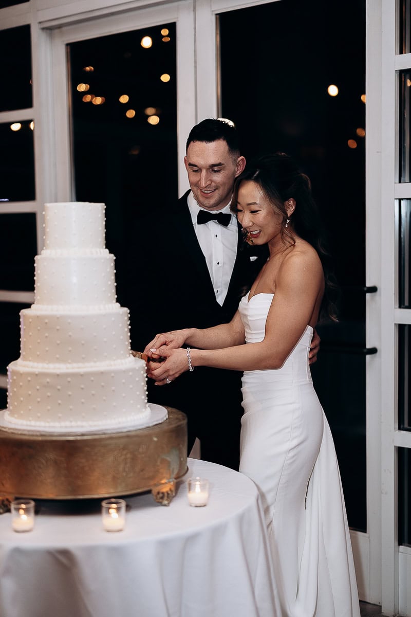Bride and groom cutting a four-tier white wedding cake on a round table with candles, inside The Stone House, Warren, NJ, at night.