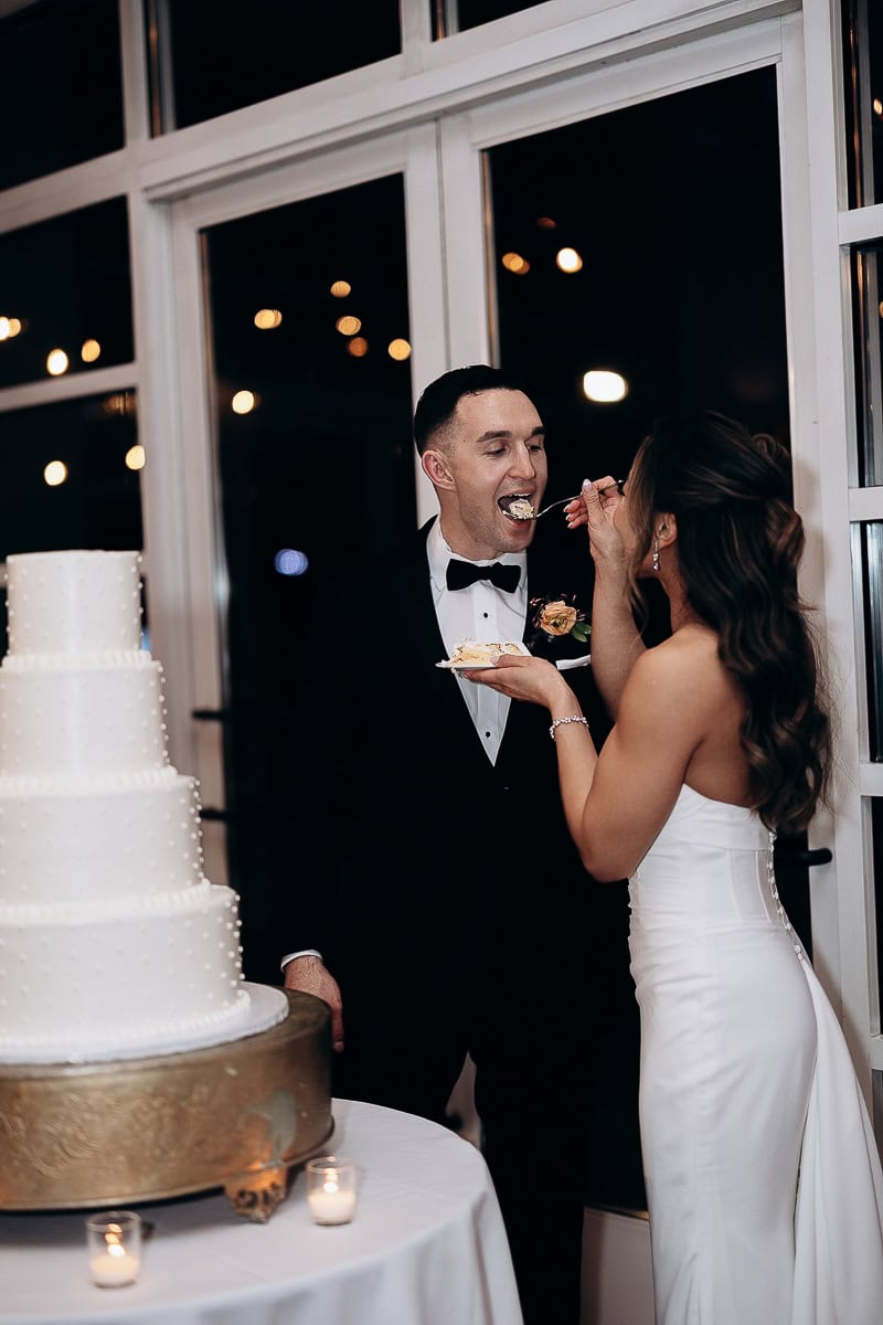 Bride in strapless white gown feeds groom in black tuxedo a bite of wedding cake beside a four-tier white cake on a gold stand with candles, indoors at night.