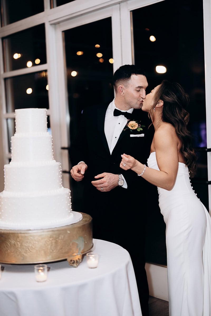 Bride and groom sharing a kiss next to a four-tiered white wedding cake on a round table with candles at The Stone House, Warren, NJ.