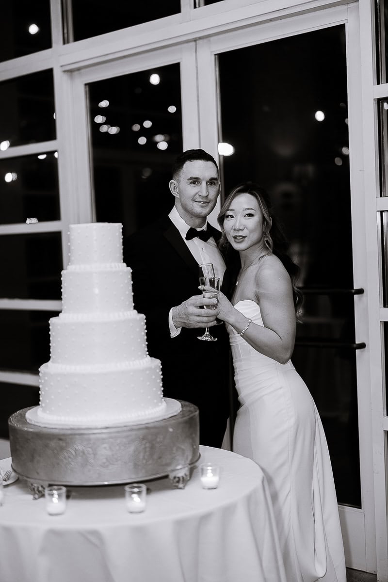Bride and groom standing beside a four-tier white wedding cake on a round table with candles, holding champagne glasses in an elegant indoor setting at night.