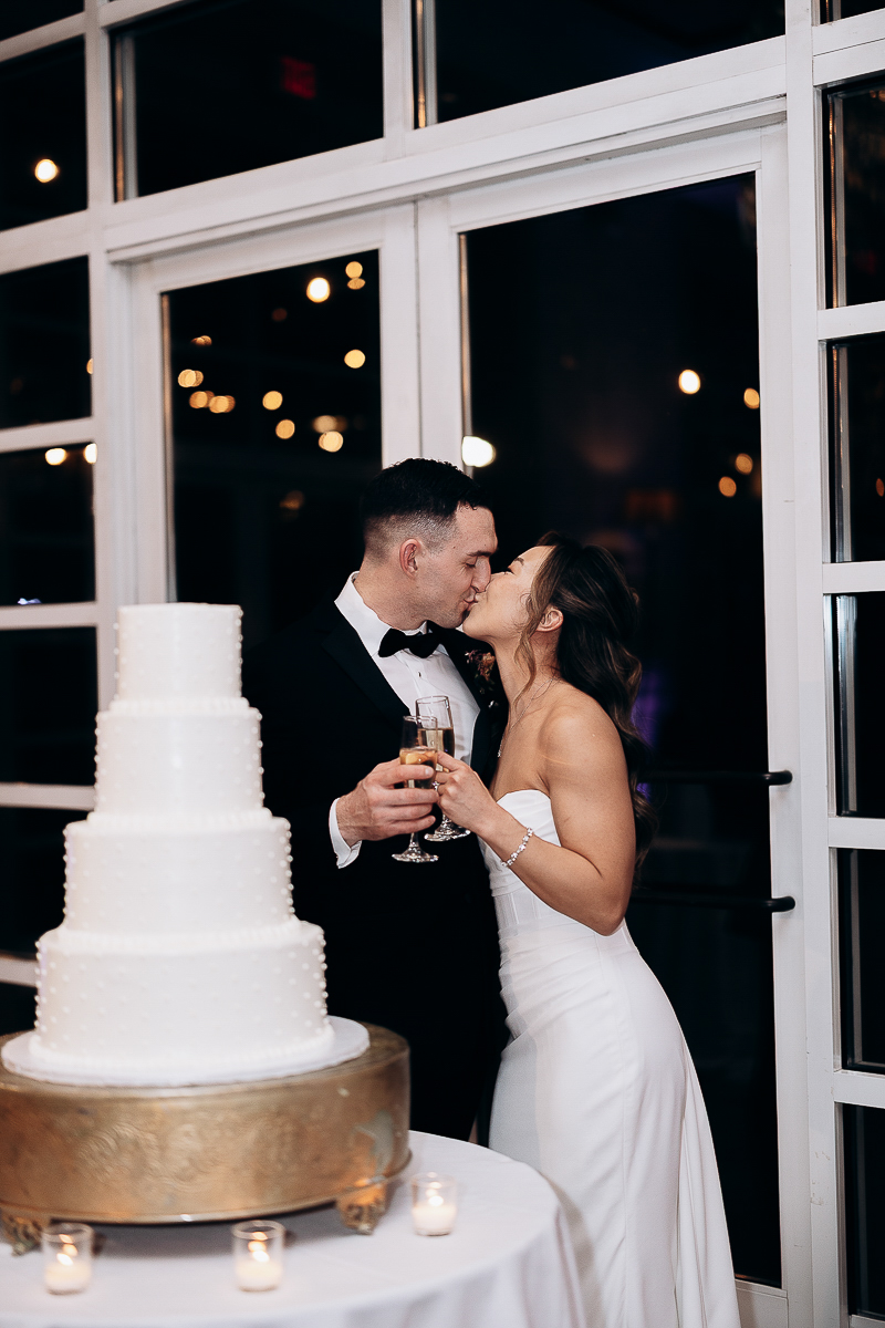 Bride and groom in elegant attire sharing a kiss while holding champagne glasses next to a four-tier white wedding cake with pearl details on a gold stand at night.