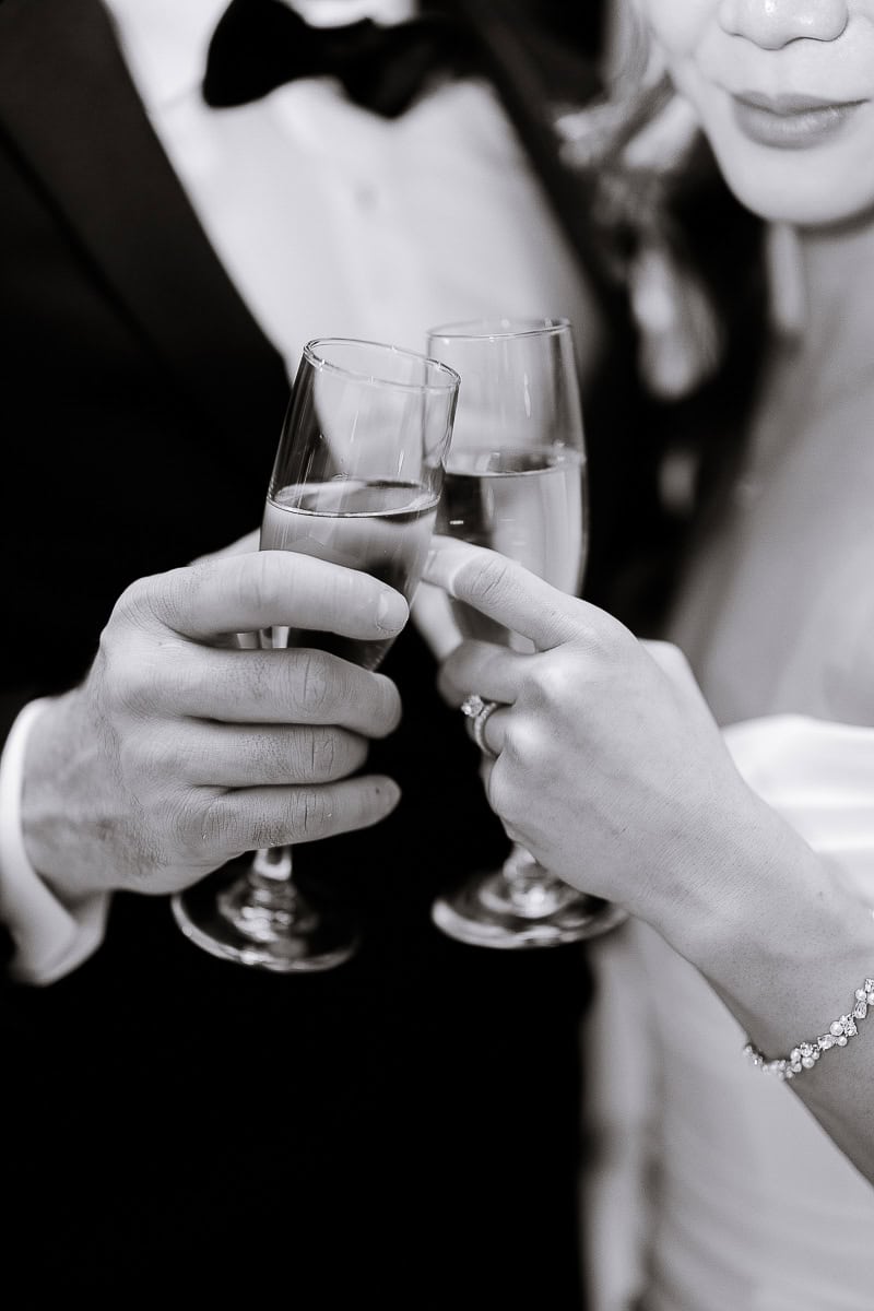 Close-up of bride and groom toasting with champagne glasses, showcasing diamond ring and bracelet, in black and white.