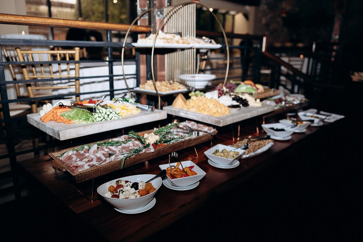 Rustic wooden table with tiered platters of sliced cured meats, fresh vegetable sticks, assorted cheeses, and small bowls of marinated vegetables and olives in a dimly lit venue.