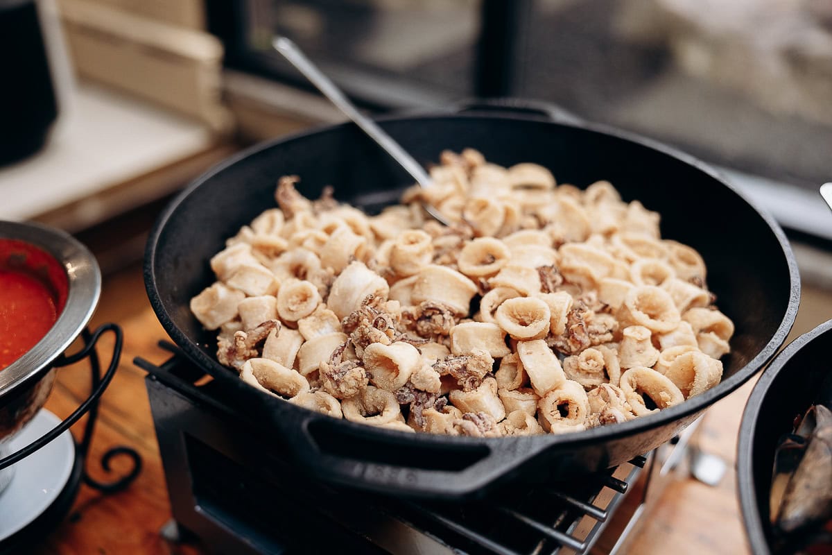 Close-up of fried calamari rings and tentacles in a black cast iron pan with a spoon, next to a bowl of red marinara sauce on a wooden table.
