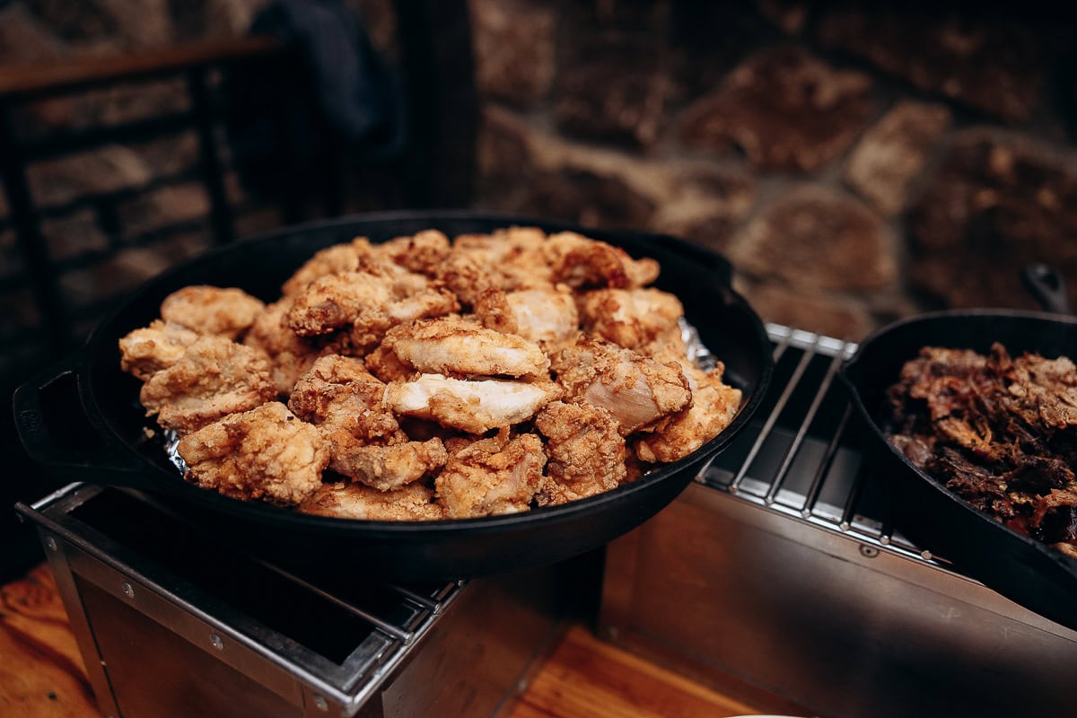 Black cast iron skillet filled with crispy fried chicken pieces on a metal warming tray with stone wall background at The Stone House, Warren, NJ.