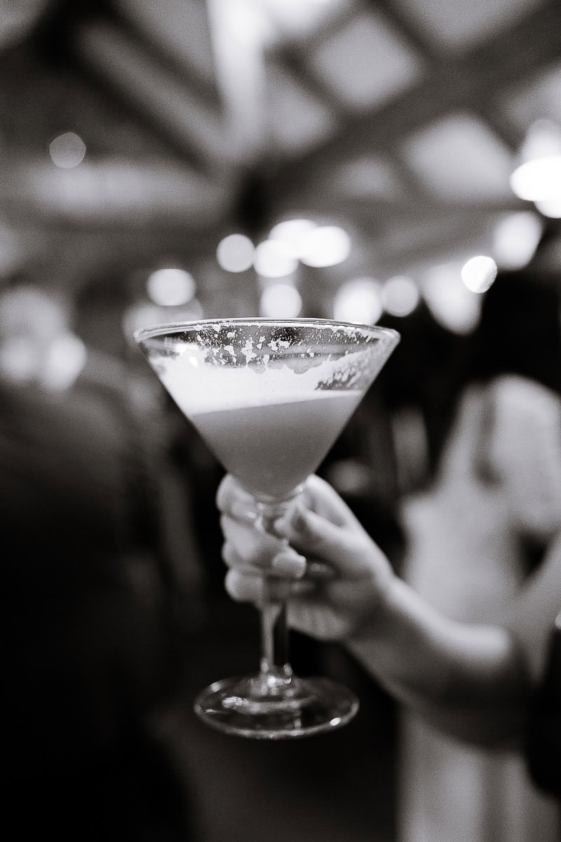 Hand holding a cocktail glass with frothy drink at a wedding reception, blurred guests and soft lighting in background.