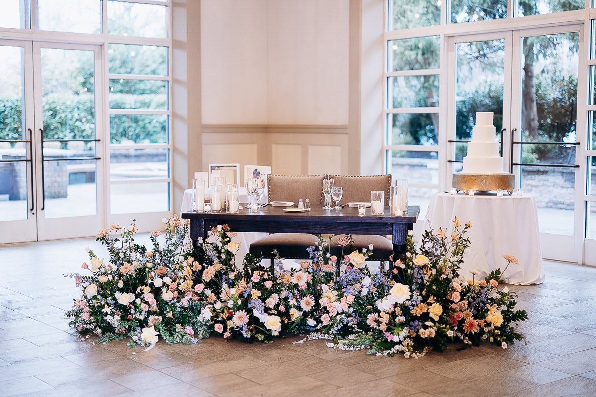 Sweetheart table with beige upholstered chairs, dark wood table adorned with glass candles and pastel floral arrangements, next to a white tiered wedding cake on a round table, inside The Stone House, Warren, NJ.