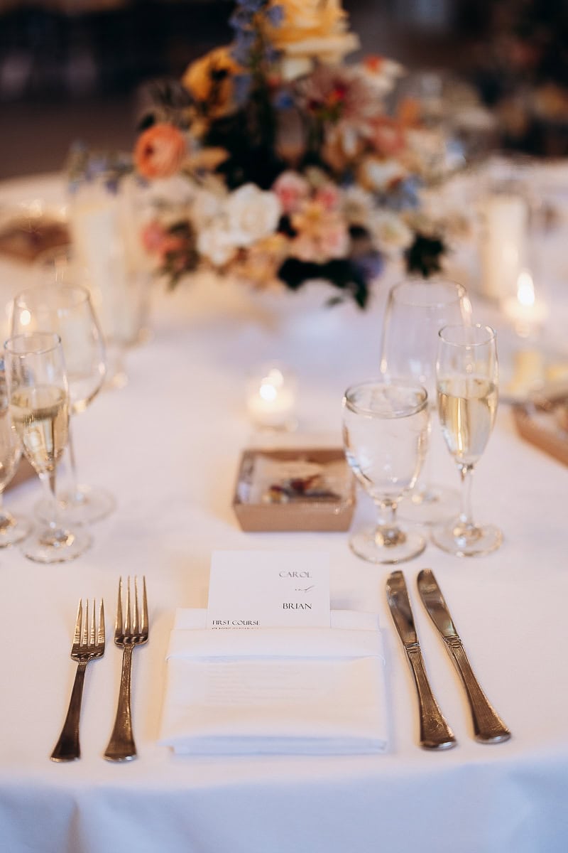 Close-up of a wedding table setting with silver forks, knives, white napkin, menu card, water and champagne glasses, and blurred floral centerpiece at The Stone House, Warren, NJ.
