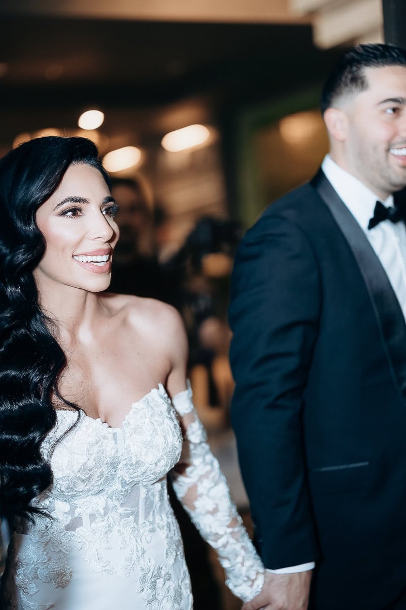 Woman in an off-the-shoulder lace wedding gown holds hands with a man in a tuxedo while walking indoors; soft bokeh lights at The Switch House, Philadelphia.