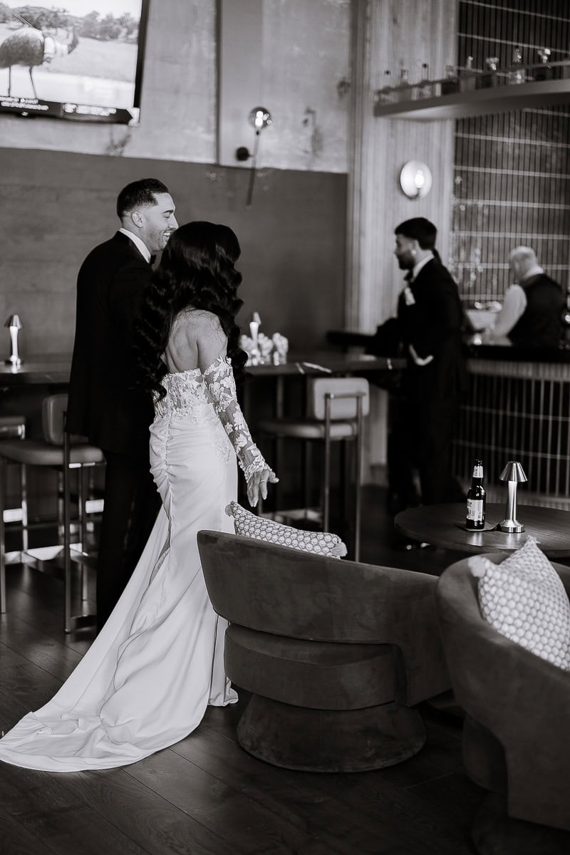 Bride in long-sleeve lace gown with silk train walks through a bar lounge beside a partner in a tux at The Switch House, Philadelphia.