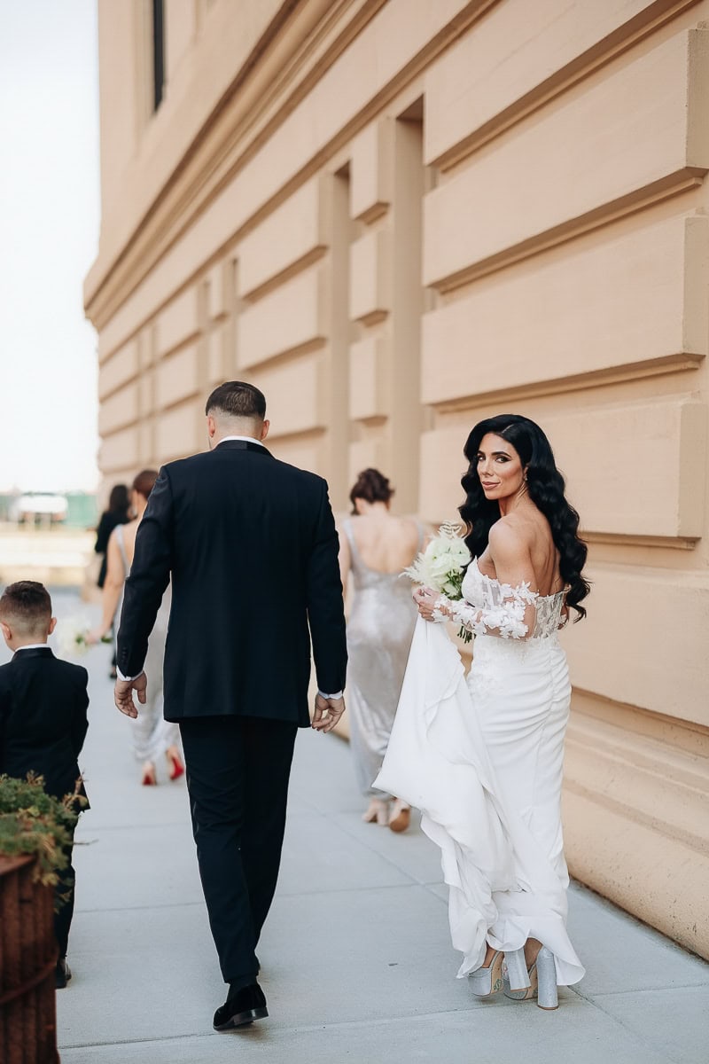 Bride in lace gown and silver platforms looks back, walking with groom along limestone facade at The Switch House by Cescaphe in Philadelphia.