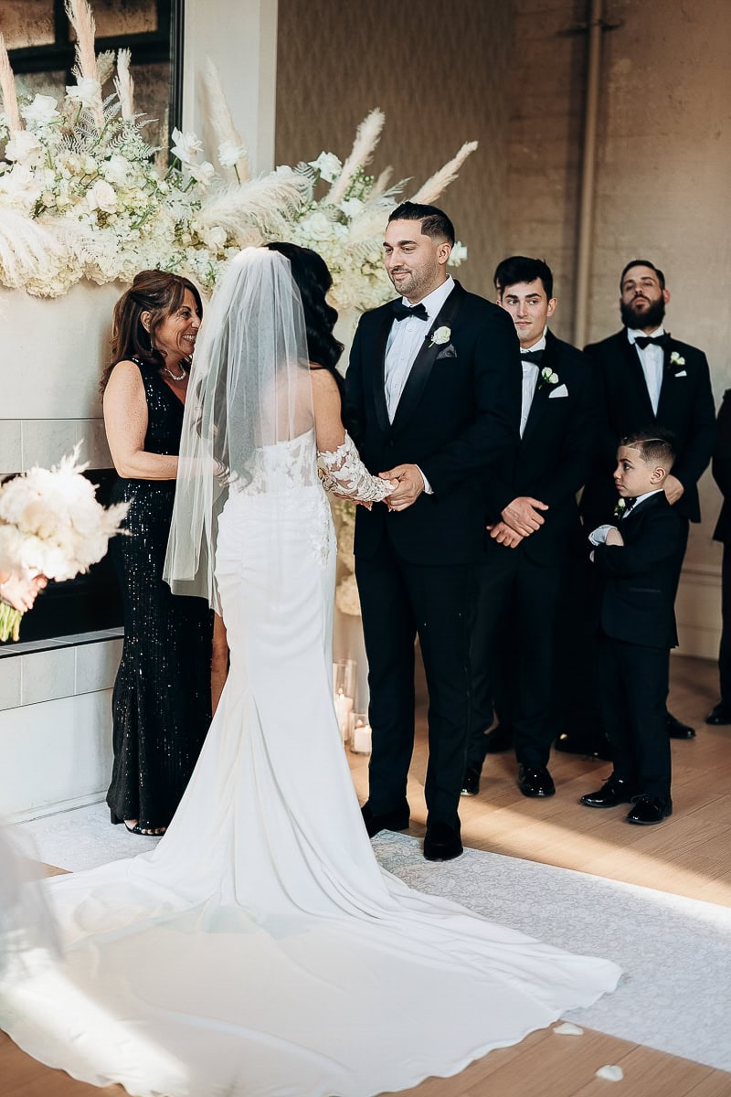 Indoor ceremony at The Switch House by Cescaphe: bride in long-sleeve gown and veil holds hands with groom before white florals and pampas.