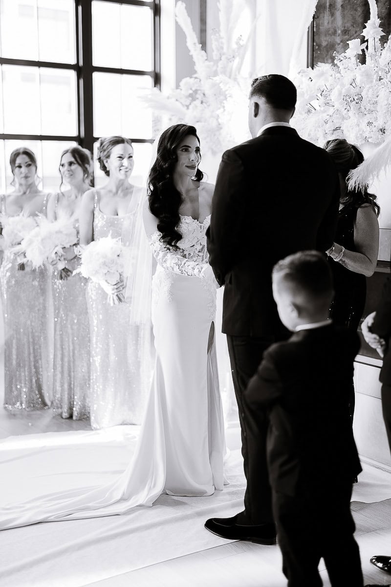 Black-and-white image of a couple at the altar, bridesmaids in sequined gowns with white bouquets; floral backdrop at The Switch House, Philadelphia.