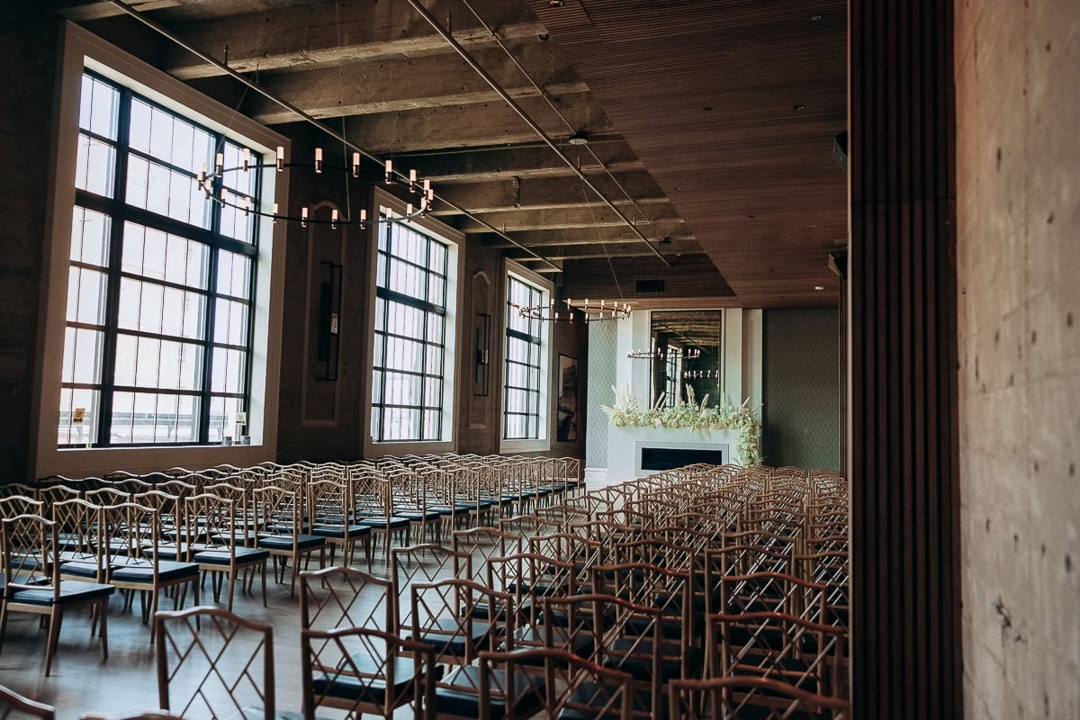 Rows of wooden lattice-back chairs face a floral-trimmed fireplace under chandeliers at The Switch House by Cescaphe, Philadelphia.