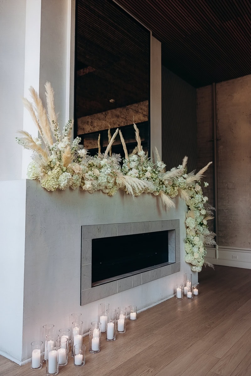 Ivory florals and pampas grass cascade over a modern fireplace with rows of glass hurricane candles at The Switch House, Philadelphia.