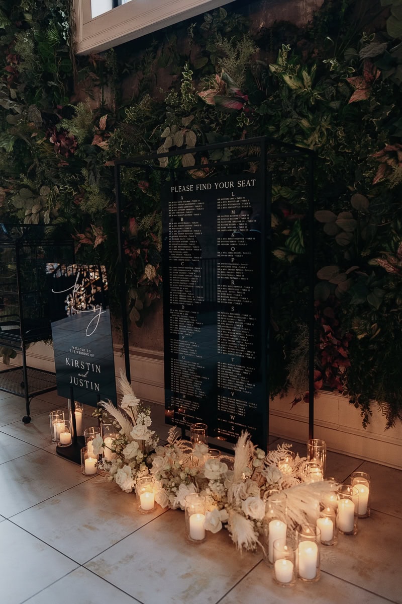Black acrylic seating chart and welcome sign before a greenery wall, surrounded by white florals and glass pillar candles at The Switch House.