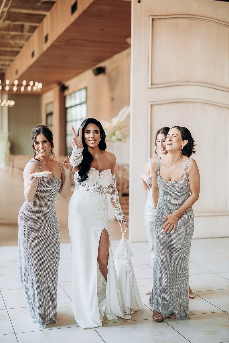 Bride in off-shoulder lace gown with thigh slit among three women in silver dresses, laughing indoors at The Switch House by Cescaphe, Philadelphia.