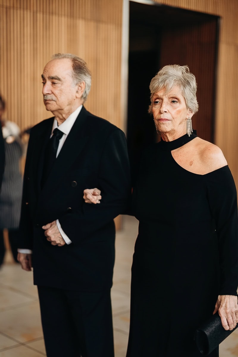Two older guests in black tie, arm in arm; woman in one-shoulder black gown with chandelier earrings and clutch, indoors at a wood‑paneled venue.