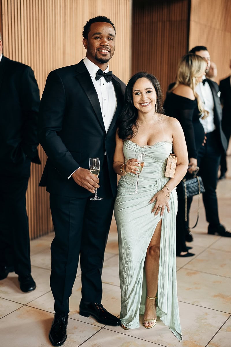 Elegant guests in black tuxedo and sage ruched gown hold champagne during cocktail hour at The Switch House by Cescaphe, Philadelphia.