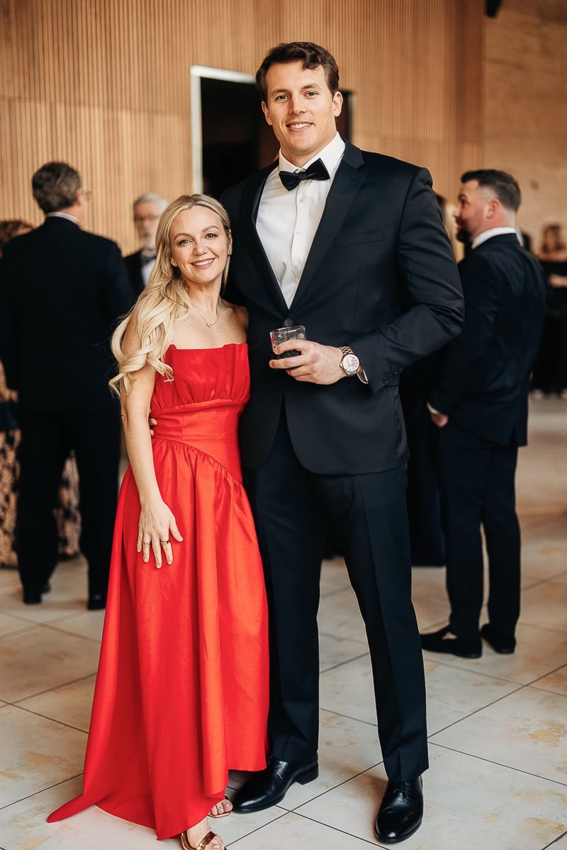 Guests pose: woman in scarlet strapless gown and gold heels beside man in black tuxedo holding a rocks glass in a wood-paneled wedding venue.