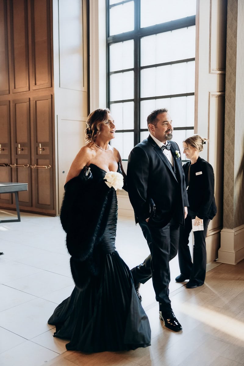 Two guests in black tie walk through sunlit hall—mermaid gown with fur wrap, man in tux. The Switch House by Cescaphe, Philadelphia.