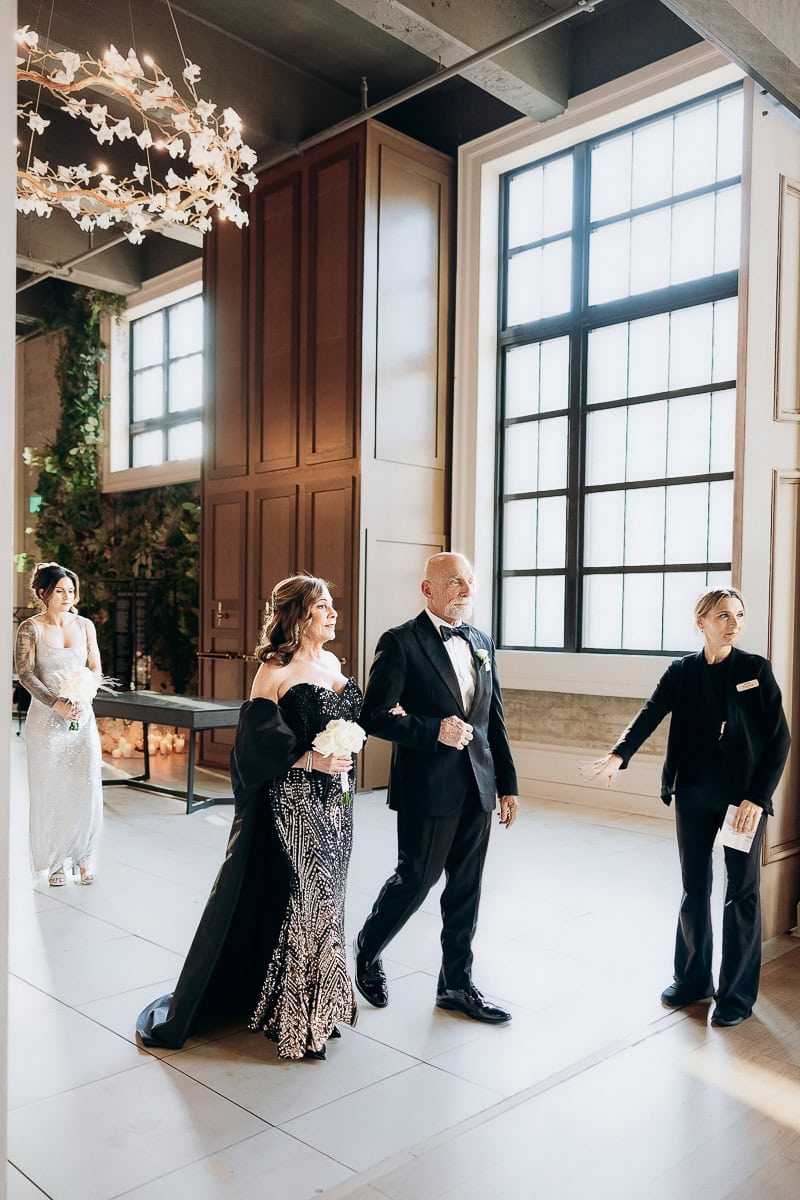 Processional in modern venue: black beaded gown and tux walk arm-in-arm with white roses, coordinator gesturing, tall grid windows beside them.