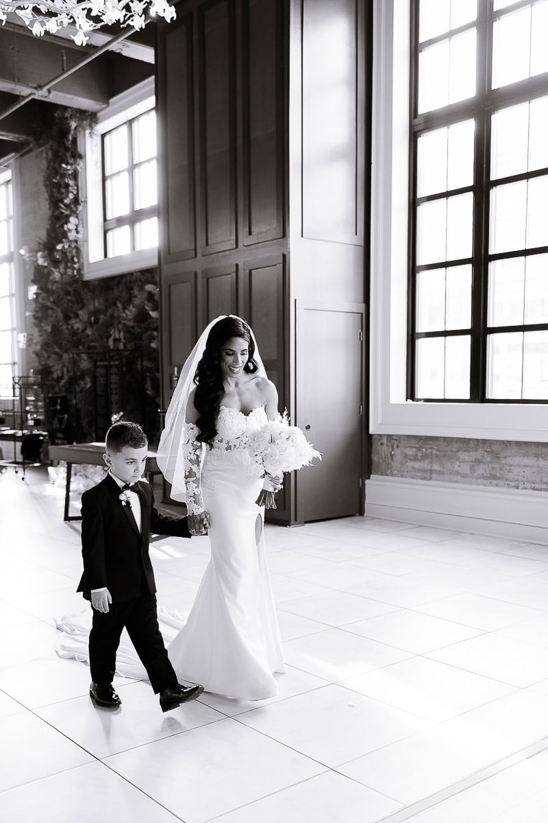 Bride in off-shoulder gown and veil walks with ring bearer, holding white bouquet, at The Switch House by Cescaphe, Philadelphia.