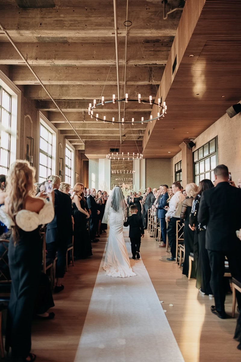 Bride and child walk down the aisle under ring chandeliers as guests stand in a window-lit loft at The Switch House, Philadelphia.