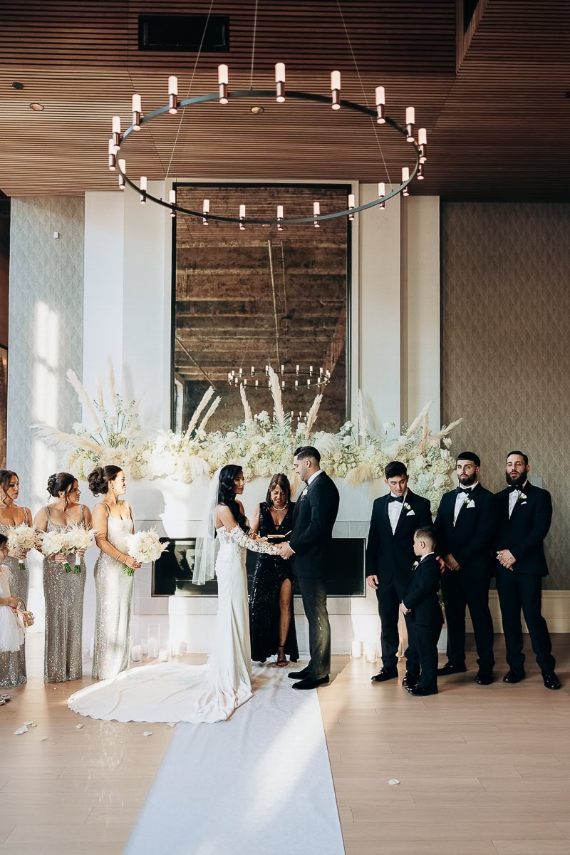 Indoor ceremony with couple exchanging vows before white florals on a fireplace, ring chandelier overhead, attendants lined on both sides, white aisle runner.
