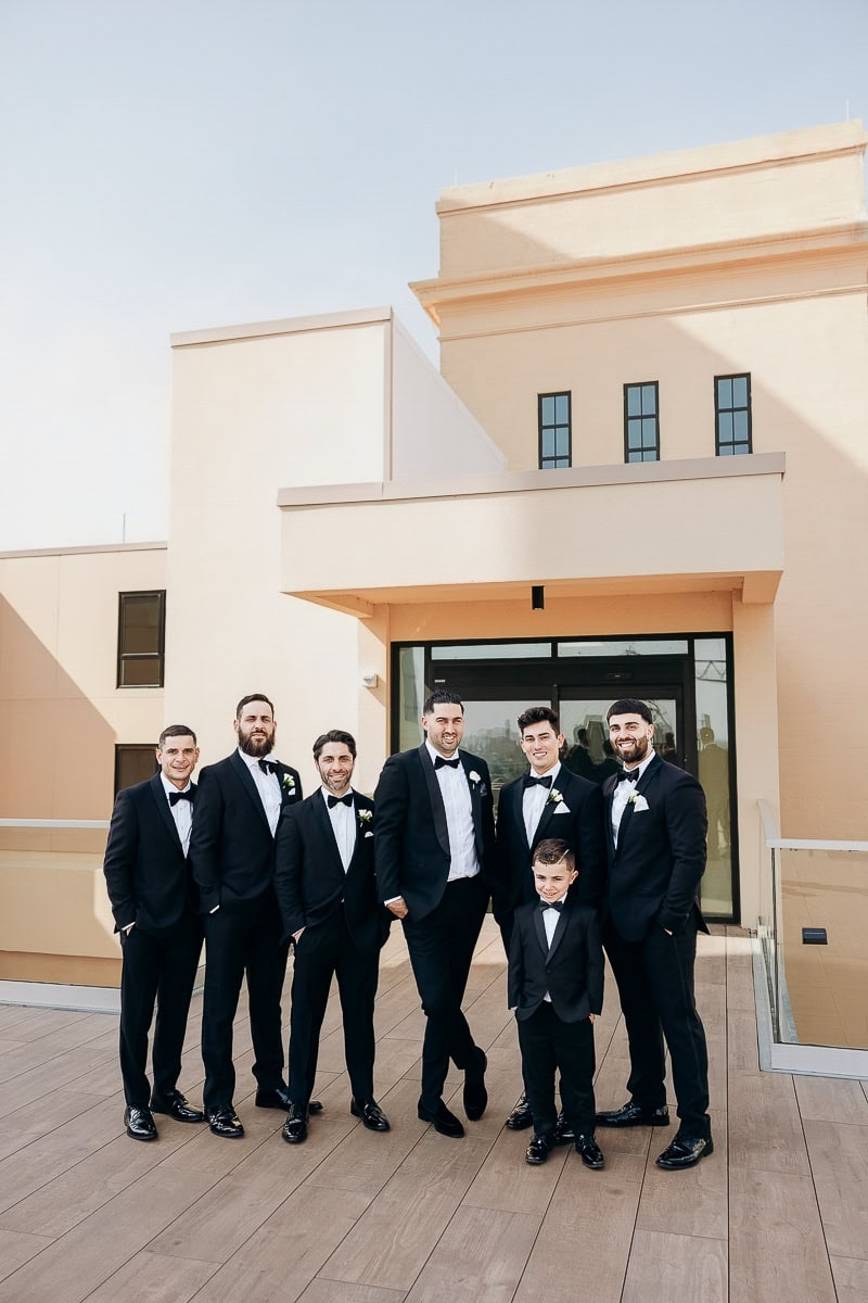 Seven men and a young boy in black tuxedos pose on a sunlit terrace with cream modern architecture at The Switch House by Cescaphe, Philadelphia, PA.